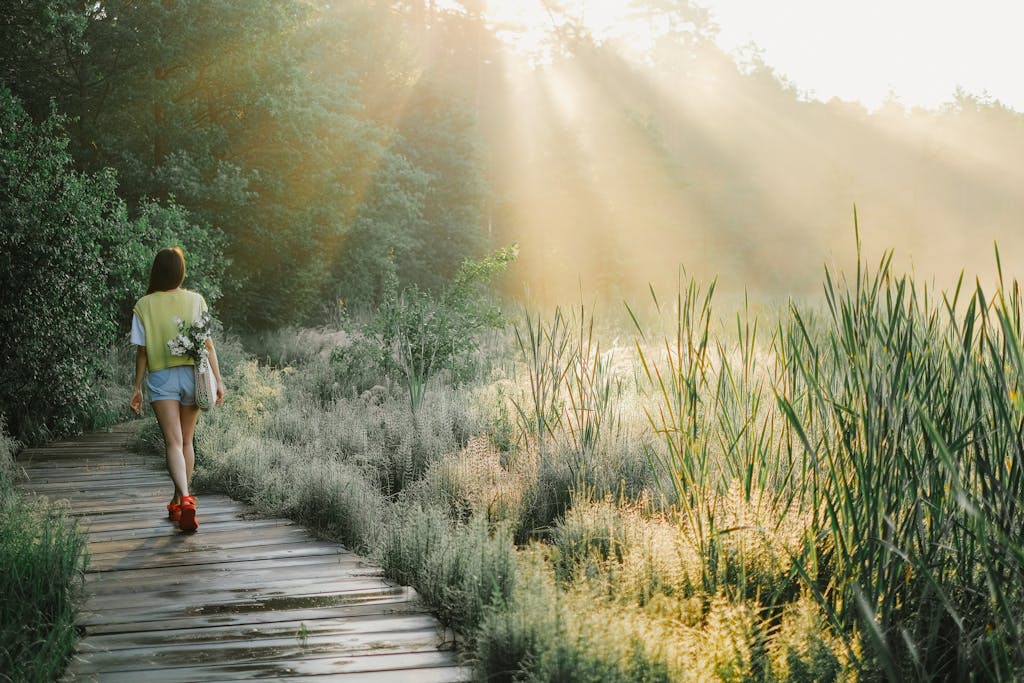 A woman enjoys a peaceful walk on a wooden path in a sunlit forest, surrounded by lush greenery.