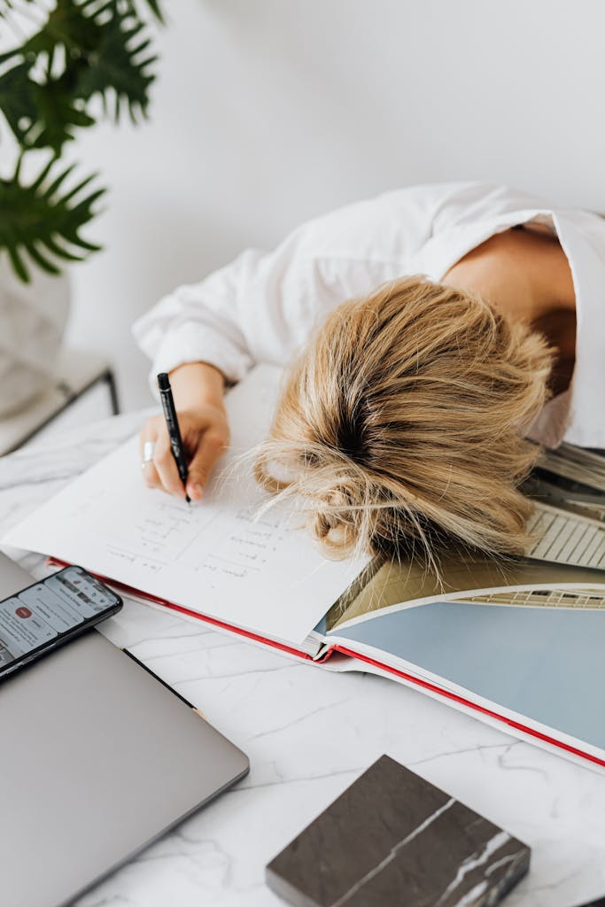 A woman expresses frustration with her head down on a desk filled with work materials in a home office setting.