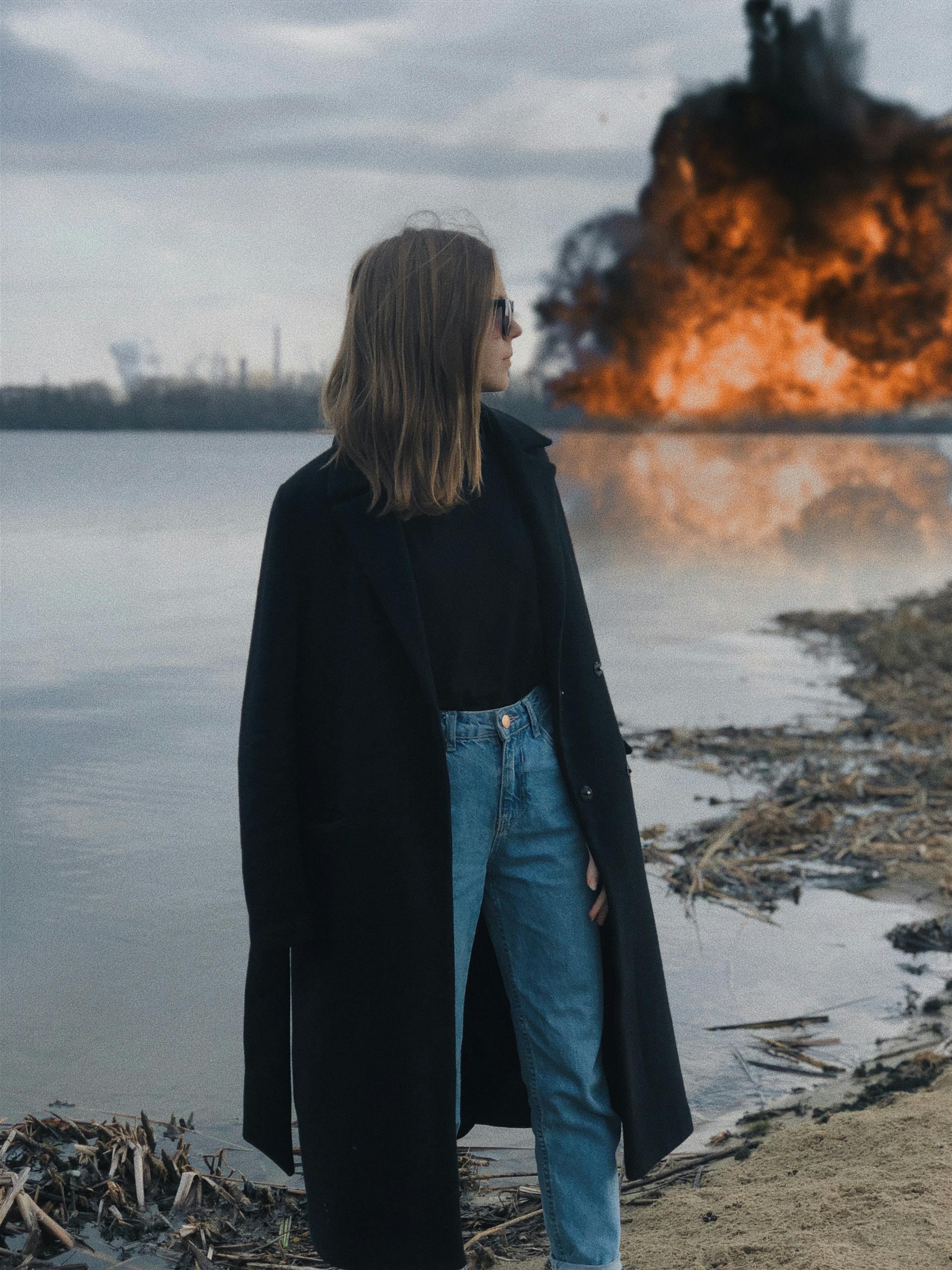 Woman in coat by lake with explosion in background, creating a dramatic scene.